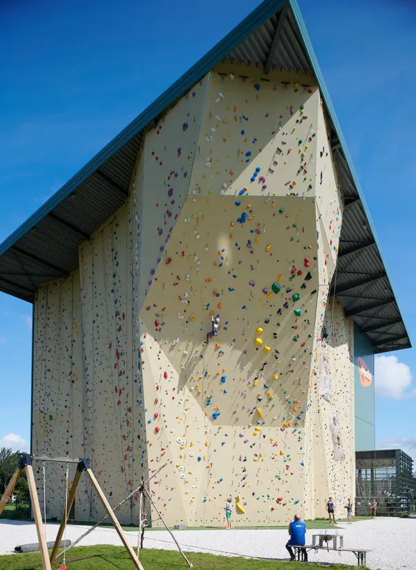 Een grote klimwand in de open lucht met kleurrijke grepen onder een schuin metalen dak, waar mensen aan het klimmen zijn en toekijken; op de achtergrond zijn een schommel en een blauwe lucht te zien.