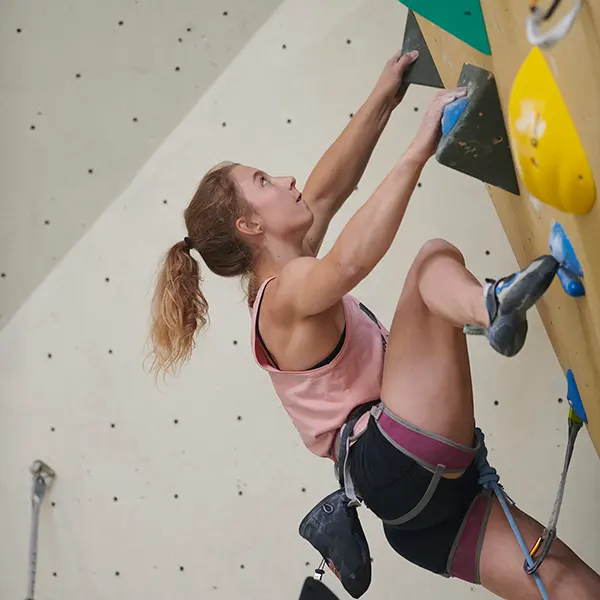 Een vrouw in een roze tanktop en klimuitrusting beklimt een indoor-klimwand; met één hand reikt ze omhoog en met de andere grijpt ze een greep vast, terwijl ze zich volledig concentreert op haar volgende beweging.