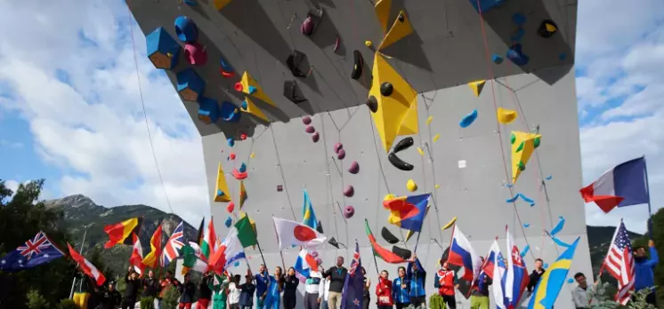 Op een zonnige dag zwaaien mensen met verschillende nationale vlaggen onder een grote klimwand in de open lucht met kleurrijke grepen, waarschijnlijk tijdens een internationale klimwedstrijd.