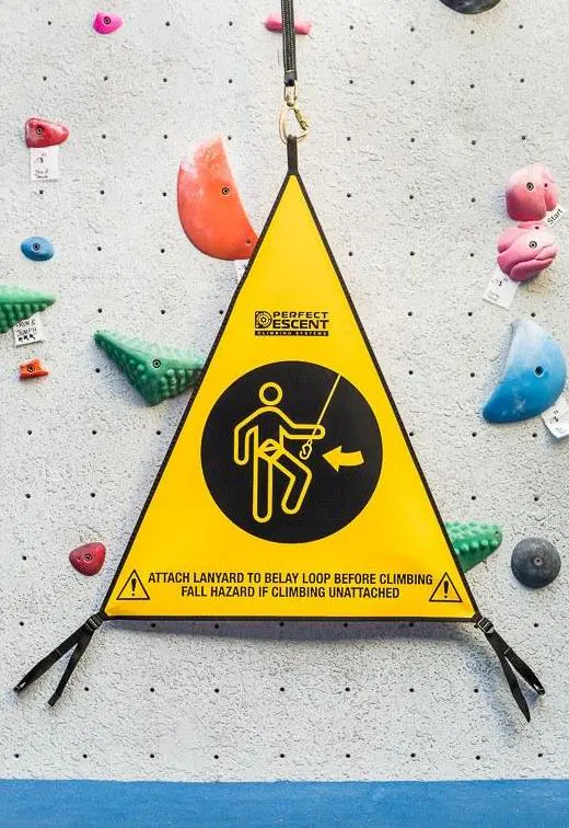 Geel driehoekig waarschuwingsbord bij de indoor-klimwand, met een afbeelding van een persoon die een veiligheidslijn aan een zekeringslus vastmaakt, en de tekst: „Maak de veiligheidslijn vast aan de zekeringslus voordat je gaat klimmen. Valgevaar als je zonder veiligheidslijn klimt.”.