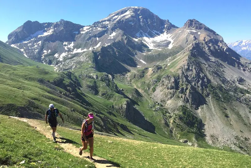 Twee wandelaars lopen over een smal pad op een met gras begroeide berghelling, met ruige, met sneeuw bestrooide bergtoppen en een helderblauwe lucht op de achtergrond.